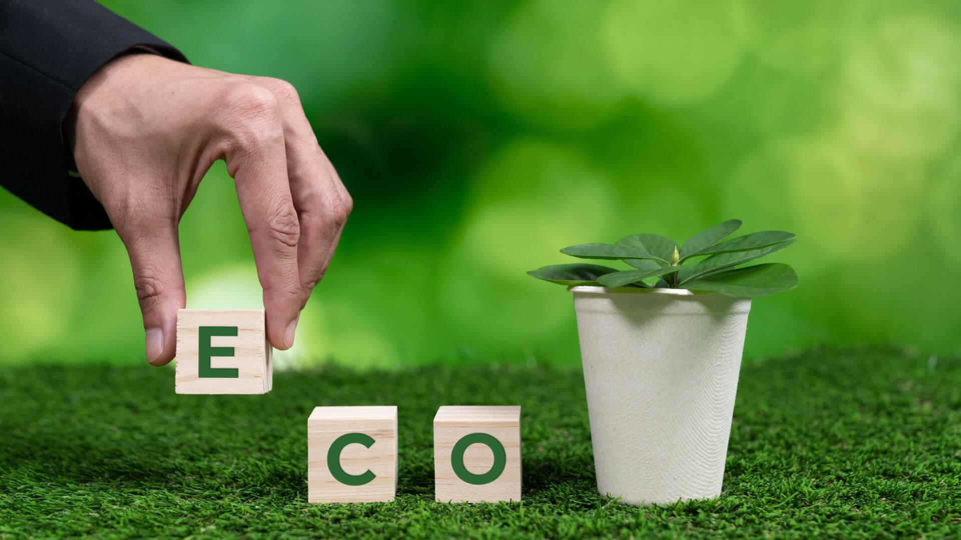 A businessman's hand placing wooden letter blocks spelling out ECO next to a plant in a sustainable cup on grass