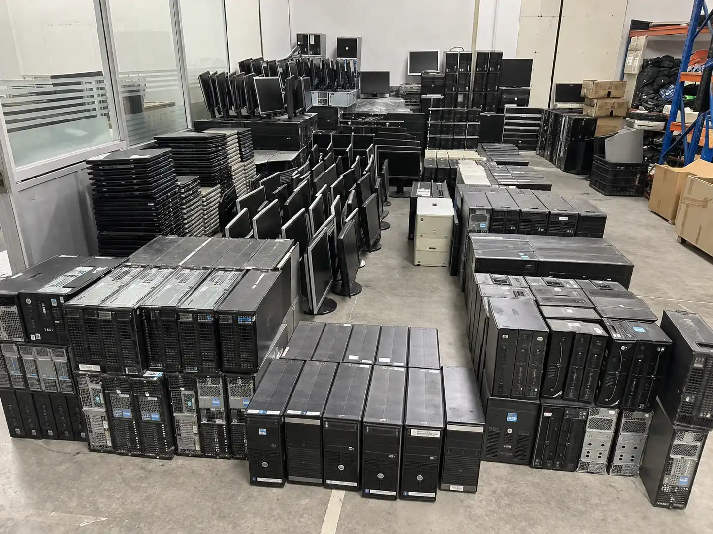 A large warehouse floor filled with rows and stacks of organized e-waste assets, including desktop tower CPUs, stacked black laptops, and computer monitors ready for hardware liquidation.