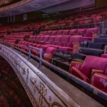 Balcony viewing deck inside a multi-tier historic theater showcasing rows of crimson seats before decorative and structural metal salvage.